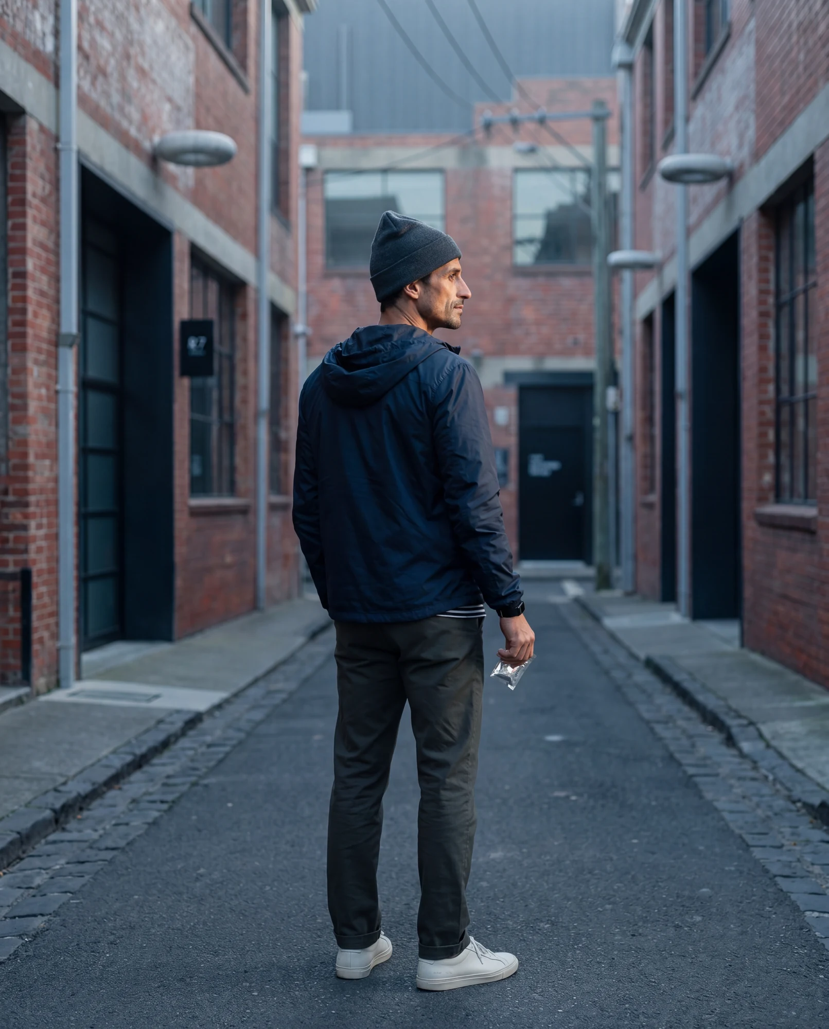 Subject MELB-01, paused in a Melbourne laneway between red brick warehouses. Navy hooded windbreaker, charcoal beanie, dark trousers, white sneakers. Right hand holds a small clear sachet.